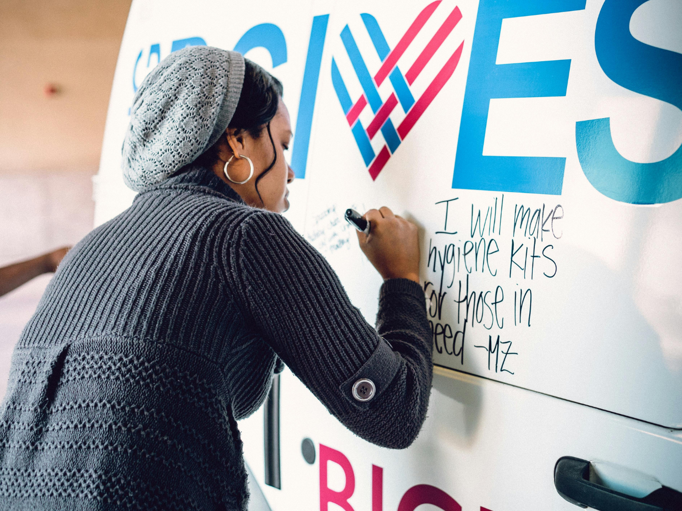 Woman writing a pledge on a Giving Tuesday campaign wall that says “I will make hygiene kits for those in need,” symbolizing community action and impact in Giving Tuesday marketing